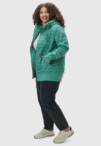 Haut à capuche en polaire verte avec un motif texturé, fermeture éclair à l'avant, capuche ajustable et poches latérales, associé à un pantalon noir et des chaussures de sport beiges.