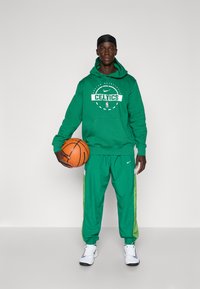 Zelená mikina Nike s logom Celtics, v kombinácii so zodpovedajúcimi zelenými tréningovými nohavicami s bledozelenými bočnými pruhmi a bielymi teniskami. Drží basketbal.