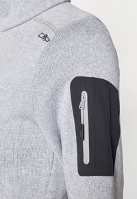 Sweatshirt en maille grise avec une finition texturée, comportant un patch en tissu noir sur le haut du bras et une poche zippée pour le rangement.