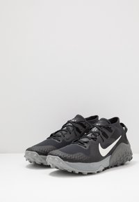 Een paar zwarte en grijze Nike trailrunning schoenen met witte swoosh-logo's en stevige zolen op een witte ondergrond.