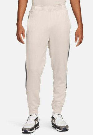 Pantalones deportivos beige con un corte tapered, cinturilla elástica, franjas laterales en gris contrastante y textura suave. Incluye bolsillos laterales.