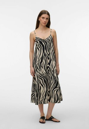 Donna in piedi che indossa un abito midi a spalline sottili con stampa zebra beige e nera e sandali neri piatti su uno sfondo chiaro e uniforme.