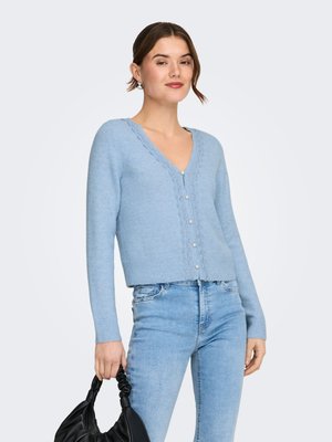 Junge Frau trägt eine hellblaue, geknöpfte Strickjacke mit Spitzenbesatz, hellblaue Jeans und hält eine schwarze, geraffte Handtasche in der linken Hand.