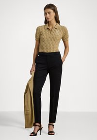 Taná pletená polo s krátkým rukávem a vzorem kabel, spárovaná s černými elegantními kalhotami a černými sandály na podpatku. Béžový blazer přehozený přes ruku.