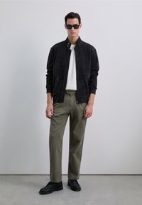 Man in zwarte suède jas, witte shirt, olijfgroene broek, zwarte schoenen en zwarte zonnebril tegen een effen lichtgrijze achtergrond.