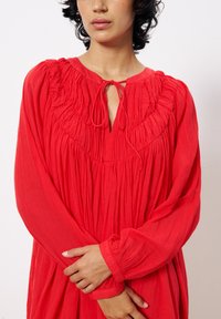 Rotes langärmliges Kleid aus leichtem Stoff mit gerafftem Ausschnitt, Bindungsdetails und plissierter Textur. Schlichte Silhouette mit sanftem Fall.