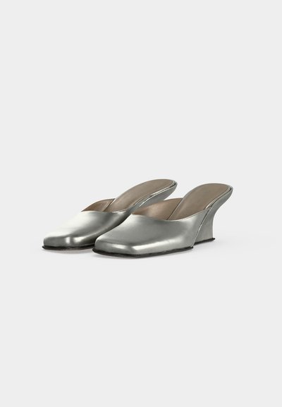 Paire de mules argentées à enfiler avec talons compensés courbés sur fond blanc uni.
