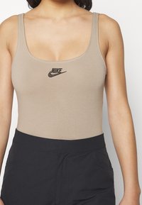 Beige ärmlös bodysuit med rund halsringning och svart Nike-logotyp i mitten. Slät tyg med minimala sömmar.
