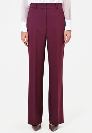 Donna che indossa pantaloni bordeaux a gamba dritta e vita alta, camicetta bianca con polsini, scarpe marroni a punta e un anello d'oro sulla mano sinistra.