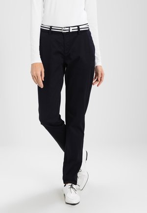 Personne portant un pantalon noir, un haut blanc à manches longues, une ceinture rayée et des chaussures blanches à lacets, debout avec une jambe croisée.