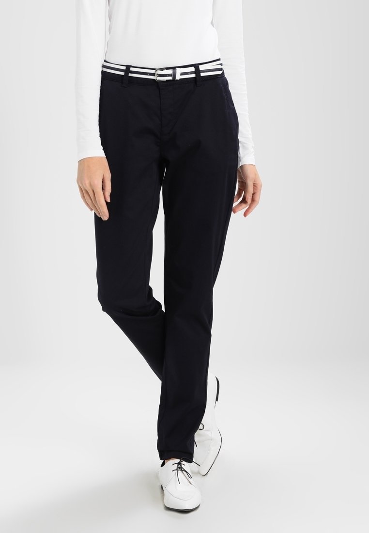 Personne portant un pantalon noir, un haut blanc à manches longues, une ceinture rayée et des chaussures blanches à lacets, debout avec une jambe croisée.