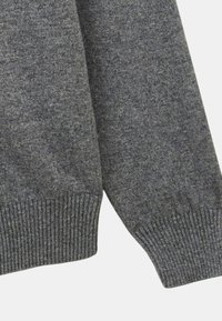 Gros plan sur un pull en tricot gris, avec des poignets côtelés et une finition texturée. Le tissu doux présente une légère variation de couleur et un design épuré.