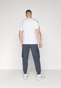 T-shirt bianco a maniche corte con dettagli neri sulle spalle, abbinato a pantaloni cargo grigio scuro con tasche laterali e scarpe sportive chiare.