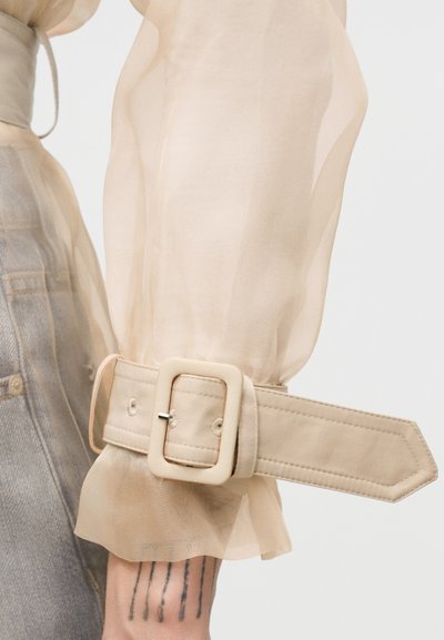 Manche de blouse beige transparente avec une grande sangle à boucle recouverte de tissu au niveau du poignet et un tatouage visible sur le bras, associée à un jean en denim clair.