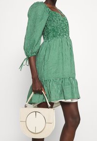 Zelené kostkované šaty s nařaseným živůtkem a vycpanými rukávy, které mají volánkový lem. Drží krémovou kabelku s kulatým akcentem.