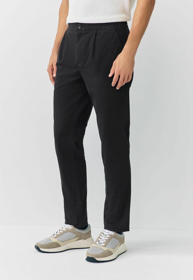 Black, lightweight trousers with a relaxed fit, featuring an elastic waistband and two side pockets. Paired with beige and gray sneakers.