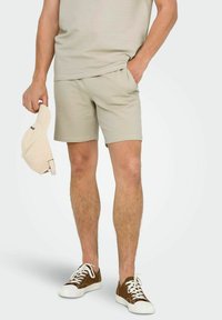 Top de tirantes texturizado beige y pantalones cortos a juego, con un corte relajado. Una gorra ligera color beige en una mano, combinada con zapatillas marrones.
