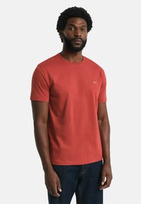 Red cotton t-shirt with a round neckline, short sleeves, and a small embroidered logo on the left side. Worn with blue denim pants.