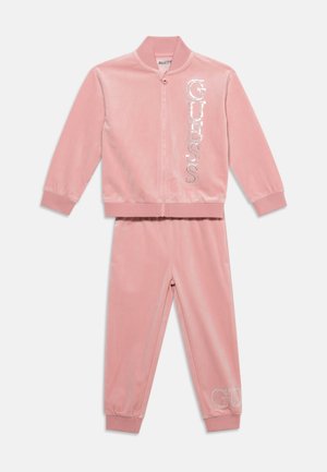Survêtement deux pièces en velours rose avec veste zippée intégrale et pantalon, avec logo "GUESS" en strass sur le devant de la veste et sur la jambe du pantalon.