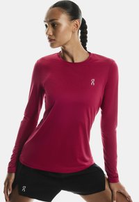 Femeie atletică cu părul împletit, îmbrăcată cu un tricou roșu cu mânecă lungă și pantaloni scurți negri, întinzându-se cu mâinile pe coapse.