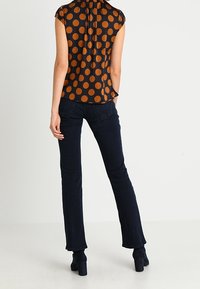 Kvinna i svart blus med stora bruna prickar och mörkblå jeans, stående med en hand i fickan, vänd bort.