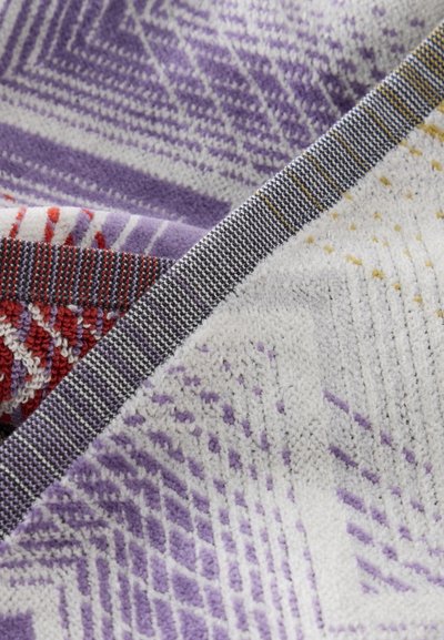Tissu plié avec des motifs géométriques complexes rouges, violets, blancs et gris, et une bordure tissée texturée.