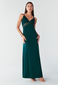 Abito maxi verde smeraldo con scollo a V, dettagli in pizzo sul corpetto e spalline sottili, realizzato in tessuto liscio e fluido.