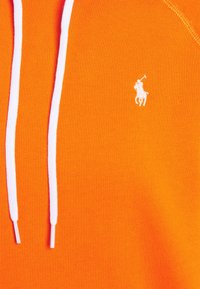 Oranžová mikina vyrobená z měkké tkaniny, která má bílé tkaničky a malé bílé vyšívané logo polo hráče na hrudi.