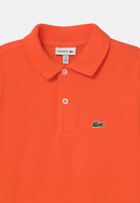 Oranžová polo košile vyrobená z texturované tkaniny, která má tříknoflíkovou légu a malé zelené logo krokodýla na hrudi.