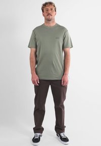 T-shirt en coton vert olive avec un col rond et un petit logo ; associé à un pantalon marron et des baskets noires avec des semelles blanches.