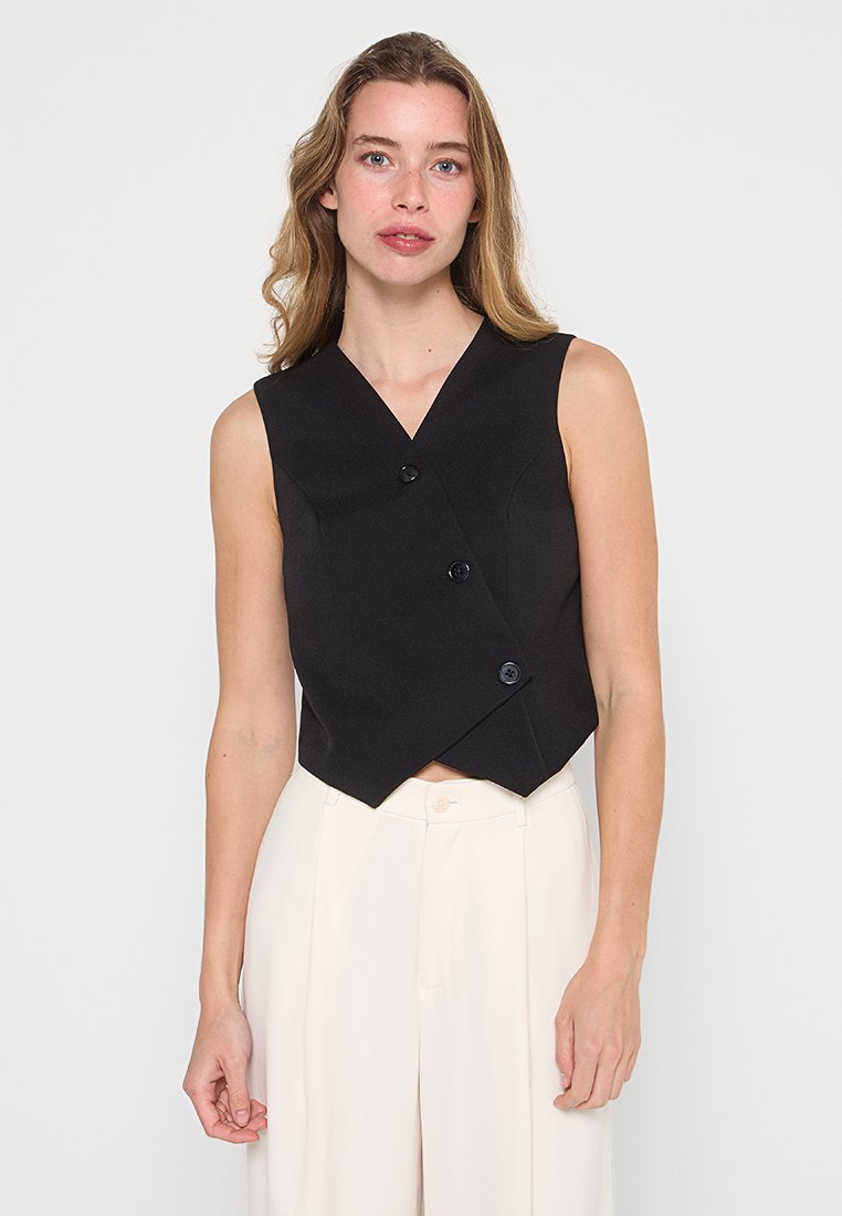 Gilet senza maniche di colore nero con design avvolgente e tre bottoni frontali; realizzato in un tessuto liscio; abbinato a pantaloni chiari.