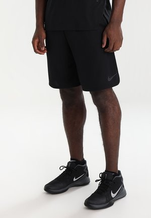 Mann trägt schwarze Nike-Shorts und schwarze Nike High-Top-Sneaker und steht auf einer weißen Oberfläche.