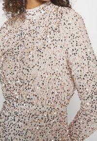 Glittrig, lätt topp med en ljus bakgrund och spridda rosaguld- och mörka paljetter, långa ärmar och subtil plisserad textur.