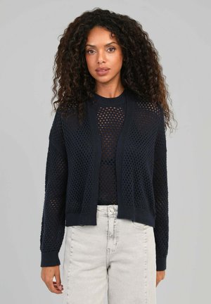 Femme aux cheveux bouclés portant un pull noir en maille ajourée et un jean taille haute gris clair, debout devant un fond gris uni.