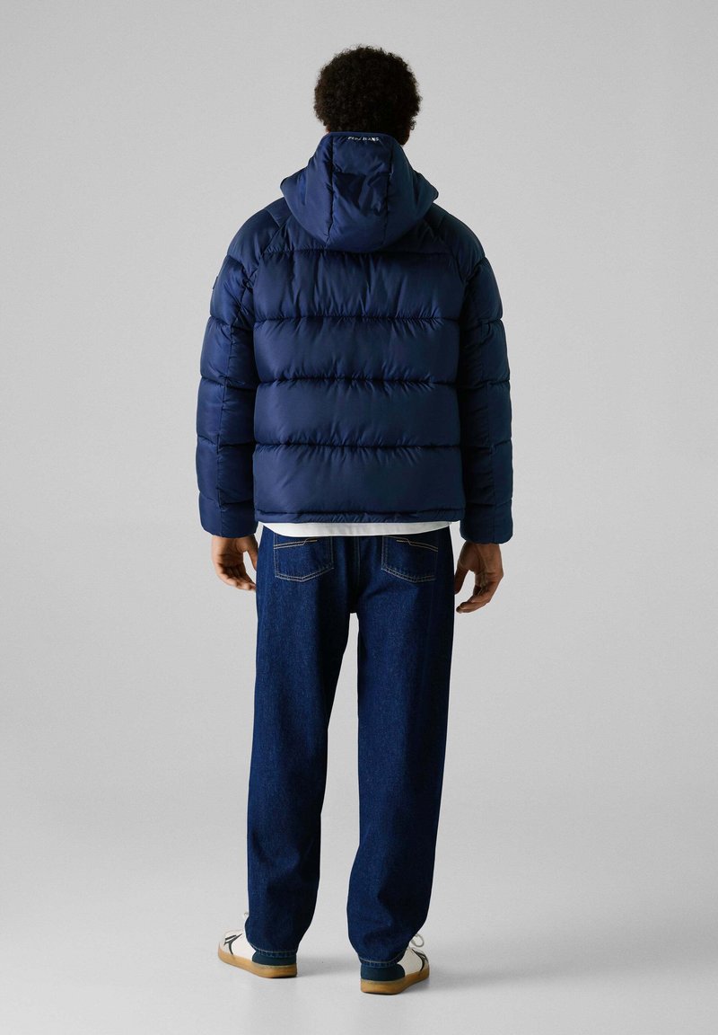 Marineblauwe gewatteerde jas met een capuchon, met een gequilt ontwerp en elastische manchetten, gedragen over een witte shirt en gecombineerd met blauwe denim.