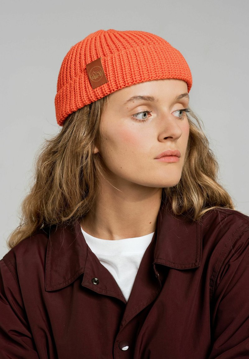 Dzianinowa pomarańczowa czapka z prążkowanym wzorem i brązową skórzaną naszywką, noszona z ciemną kurtką i białą koszulą.