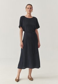 Donna con i capelli pettinati all'indietro che indossa un vestito midi nero texturizzato con maniche corte, una cintura in vita, orecchini a cerchio d'oro e décolleté marroni con punta.