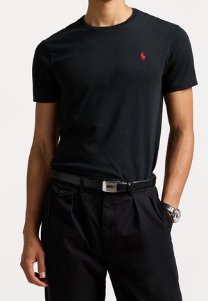 Man draagt een nauwsluitend zwart T-shirt met een klein rood logo op de borst, een zwarte riem, zwarte broek en een zilveren polshorloge aan de linkerpols.