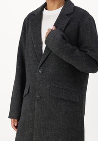 Uhlově šedý vlněný kabát s klopovým límcem, dvěma předními kapsami a dvěma knoflíky. Materiál má texturovaný povrch.