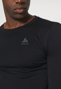 Črna majica z dolgimi rokavi, izdelana iz gladkega materiala, z zaobljenim izrezom in eleganco prileganjem, ter subtilnim sivo-odlo logotipom na prsih.