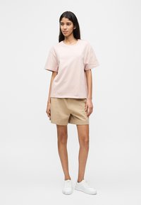 Νεαρή γυναίκα στέκεται φορώντας ένα ανοιχτό ροζ κοντομάνικο t-shirt, μπεζ σορτς και λευκά αθλητικά παπούτσια μπροστά σε απλό φόντο.