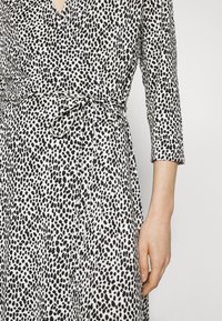 Schwarz-weißes Wickelkleid mit gepunktetem Muster, dreiviertellangen Ärmeln und einer gebundenen Taillendetail. Glatte Stofftextur und fließendes Design.