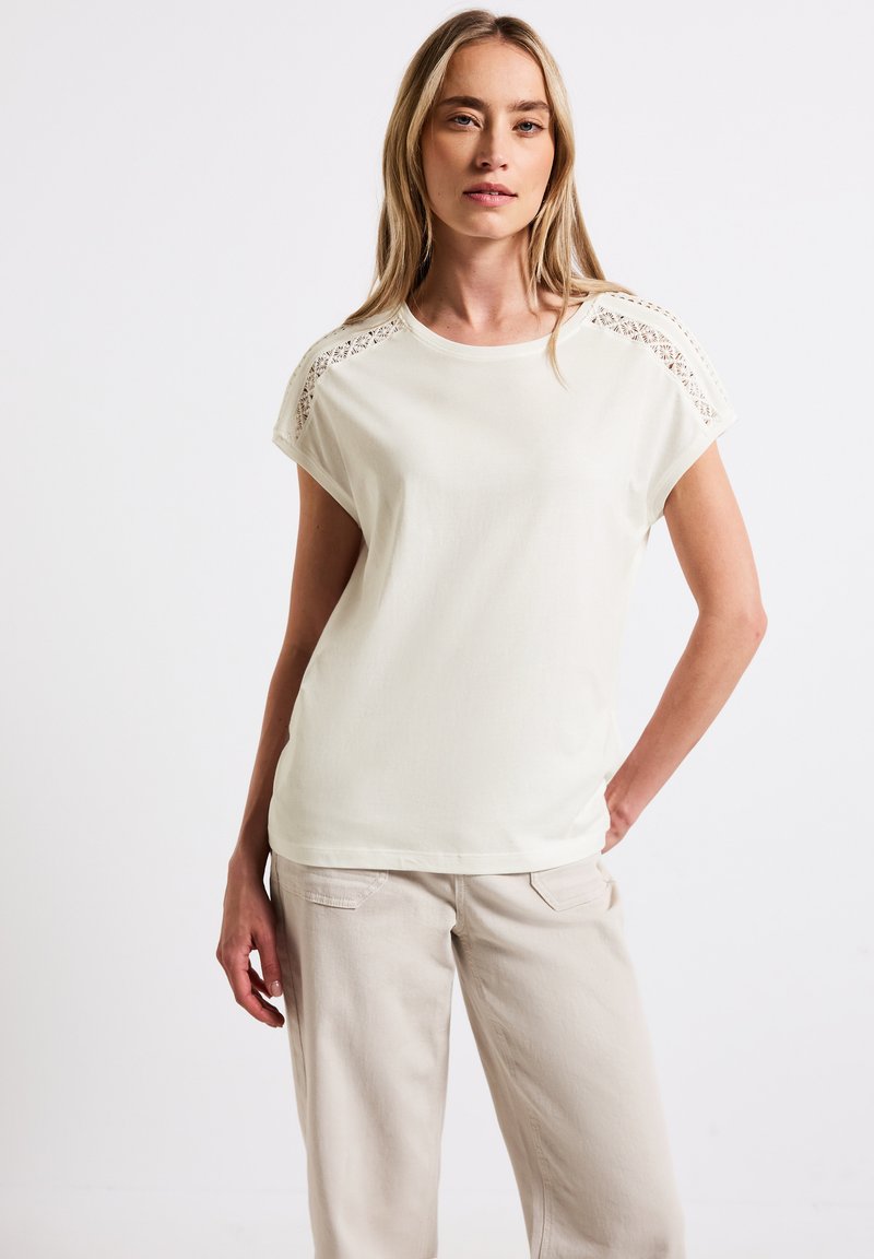 Creme kortærmet t-shirt med blonde detaljer på skuldrene, med rund hals og afslappet pasform, parret med lyse beige bukser.
