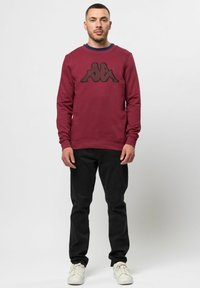 Burgunderfarget hettegenser med svart logo-design på forsiden. Rund hals og ribbestrikkede mansjetter, sammen med svarte bukser og hvite sneakers.
