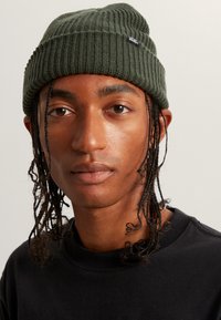 Gorra de punto acanalado en verde oliva con una etiqueta de marca Vans negra, que presenta un ajuste ceñido y una textura de tela. Combinada con una sudadera negra.