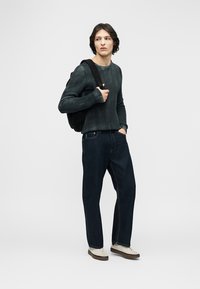 Νεαρός άνδρας με σκούρο πράσινο πουλόβερ, σκούρο τζιν, λευκά αθλητικά παπούτσια και μαύρο σακίδιο, στέκεται με το ένα χέρι στην τσέπη, κοιτάζοντας πλάγια.