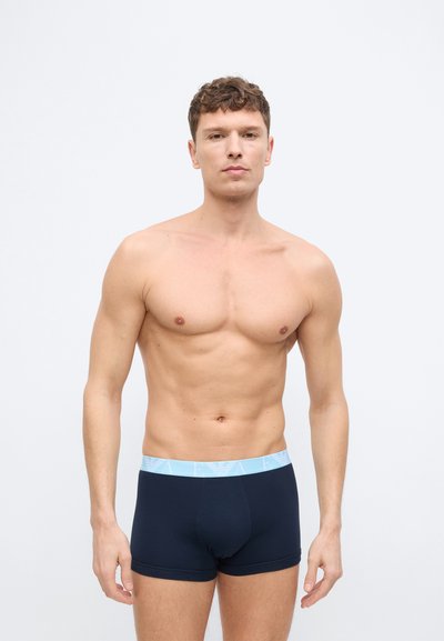 Νεαρός άνδρας με κοντά καστανά μαλλιά, φορώντας μπλε μποξεράκια με ανοιχτό γαλάζια ζώνη μέσης, στέκεται μπροστά σε απλό ανοιχτό γκρι φόντο.