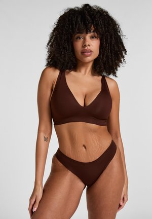 Frau mit lockigem Haar, die ein braunes Bikini-Set trägt, steht vor einem neutralen Hintergrund mit einem Schmetterlings-Tattoo am Arm.