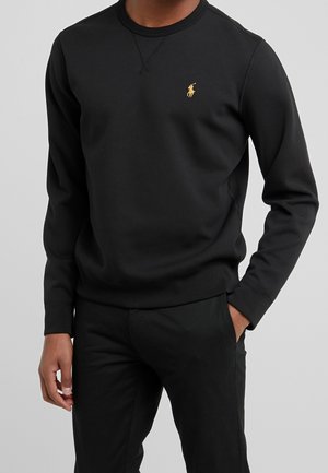 Uomo che indossa una felpa nera a maniche lunghe con un piccolo logo giallo di un giocatore di polo sul petto e pantaloni neri.