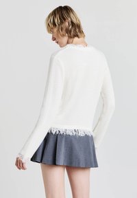 Maglione bianco strutturato con orlo sfrangiato, maniche lunghe e vestibilità casual, abbinato a una gonna svasata grigia scura. Vista dal retro, dettagli minimi visibili.